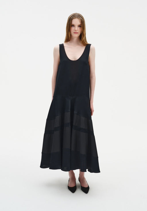 Front view of a model wearing a sleeveless black midi dress with a scoop neckline and a softly flared, semi-sheer panelled skirt, paired with black pointed shoes.