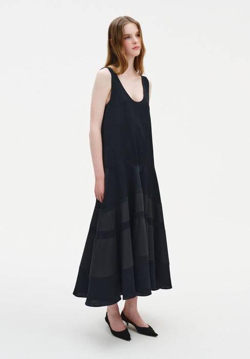 Side view of a model wearing a sleeveless black midi dress with a scoop neckline and a softly flared, semi-sheer panelled skirt, paired with black pointed shoes.
