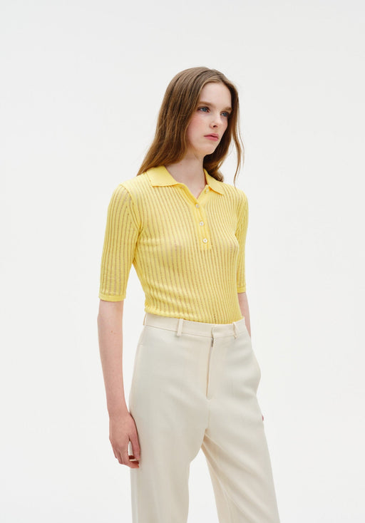 Three-quarter view of a model wearing a fitted yellow ribbed short-sleeve polo top with buttons, paired with high-waisted cream trousers.