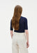 A woman with long light brown hair is shown from the back in a relaxed standing pose against a plain background, with her head slightly turned and her posture upright.