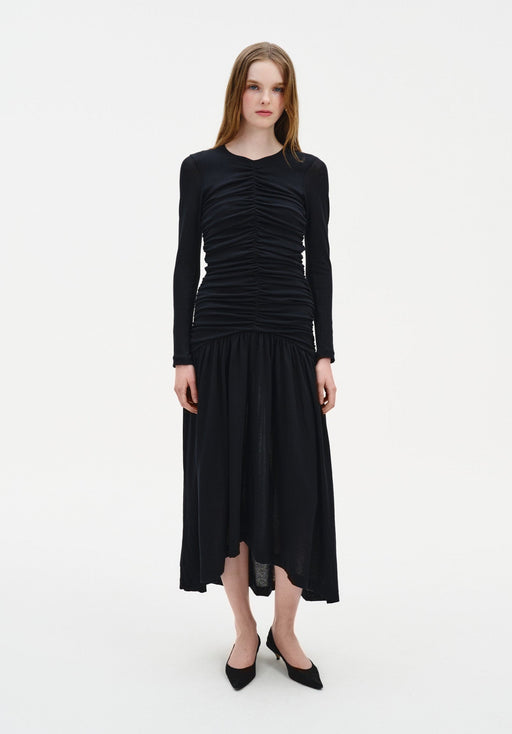 A woman stands facing forward against a plain white background, wearing a black long-sleeve dress with ruched detailing on the bodice and a loose, mid-calf skirt. She has straight, light brown hair worn down and is dressed with black pointed-toe heels, maintaining a neutral expression with arms relaxed at her sides.