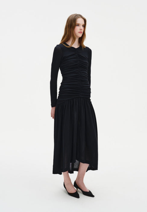 A young woman with long light brown hair stands against a plain white background, wearing a fitted black long-sleeve dress with ruched detailing and a flowy mid-calf skirt, paired with black pointed heels. She faces slightly to the side with a neutral expression and relaxed posture.