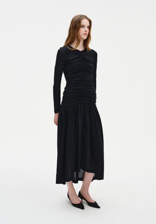 A young woman with long light brown hair stands against a plain white background, wearing a fitted black long-sleeve dress with ruched detailing and a flowy mid-calf skirt, paired with black pointed heels. She faces slightly to the side with a neutral expression and relaxed posture.