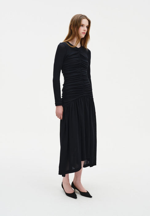 A woman stands in a three-quarter side pose against a plain white background, wearing a fitted black long-sleeve dress with ruched detailing along the front and a flowy mid-calf skirt, paired with black pointed heels. Her straight light brown hair is worn down, and she looks slightly off to the side with a neutral expression.
