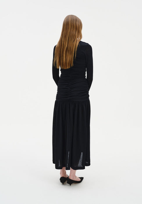 Rear view of a woman standing against a plain white background, wearing a fitted black long-sleeve dress with ruched detailing through the back and a flowy mid-calf skirt. Her straight light brown hair falls down her back, and she wears black pointed heels.