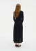 Rear view of a woman standing against a plain white background, wearing a fitted black long-sleeve dress with ruched detailing through the back and a flowy mid-calf skirt. Her straight light brown hair falls down her back, and she wears black pointed heels.