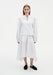 Front view of a woman wearing a crisp white long-sleeve top with a notched neckline and structured shoulders, paired with a white asymmetrical midi skirt and black slip-on flats.