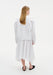 Back view of a woman wearing a white long-sleeve top with structured shoulders and a clean, minimal cut, paired with a flowing white asymmetrical midi skirt and black slip-on flats.