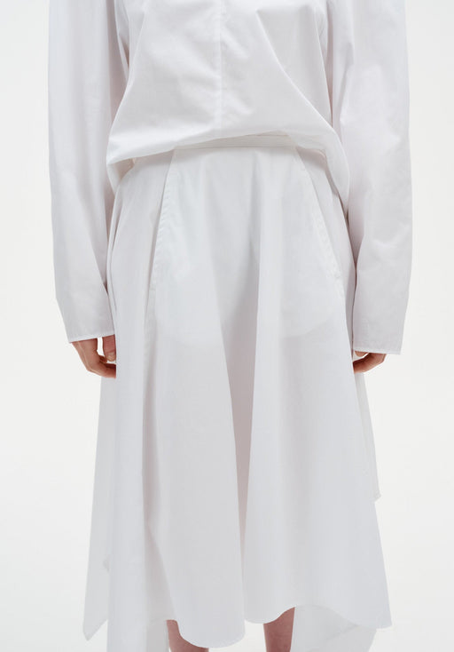 Close-up of a white outfit showing a long-sleeve top tucked into a high-waisted skirt with soft pleats and a fluid, asymmetrical hem.