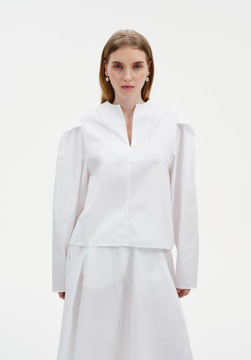 Front view of a model wearing a white blouse with a V-slit neckline and structured shoulders, paired with a matching flowing skirt, shown against a plain background.