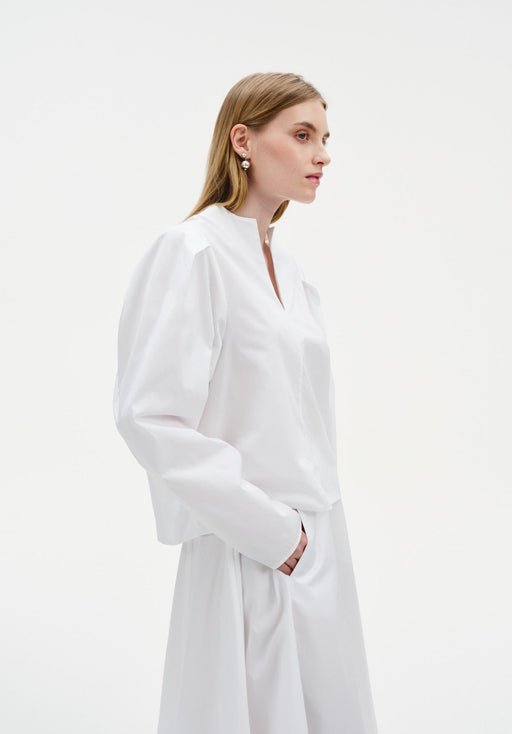 Side view of a model wearing a white blouse with a V-slit neckline and voluminous sleeves, paired with a matching flowing skirt, hands in pockets.