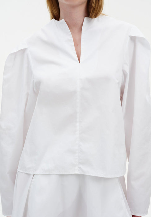 Close-up of a white blouse with a clean V-slit neckline, structured shoulders, and long sleeves in a smooth, crisp fabric.