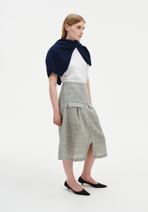 A woman stands in a three-quarter side pose against a plain white background, wearing a white top with a navy sweater draped over her shoulders, paired with a light gray textured midi skirt featuring a front slit and flap pocket detail, and black pointed heels. Her straight light brown hair is worn down, and she looks slightly to the side with a neutral expression.