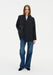 A woman with long light brown hair stands facing forward against a plain background, posing with a relaxed stance, one hand in her pocket, and a neutral expression.