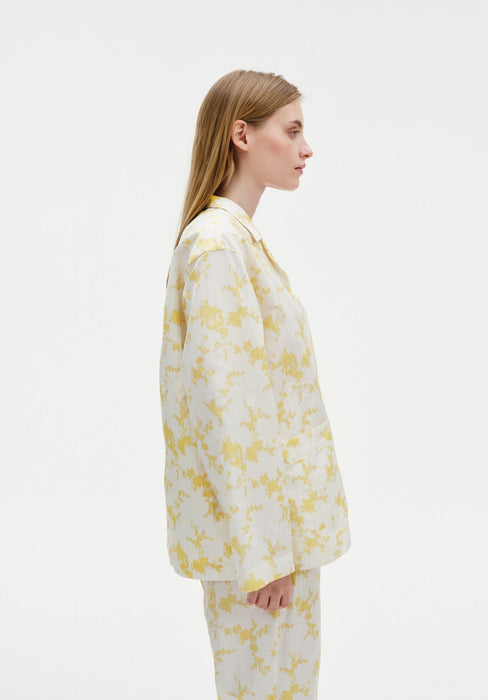Side profile of a woman wearing a white long-sleeve shirt and matching trousers with a soft yellow floral pattern, highlighting the relaxed fit and clean silhouette against a plain studio background.
