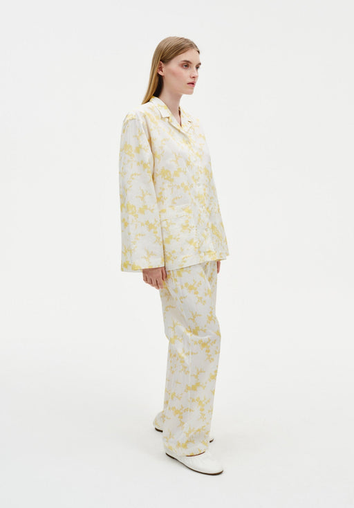 Side view of a model wearing a cream two-piece set with a yellow abstract floral print, featuring a long-sleeve collared jacket-style top and relaxed straight-leg trousers, paired with white shoes.