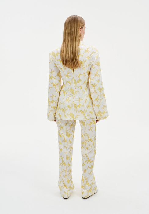 Back view of a model wearing a cream two-piece outfit with a yellow abstract floral print, featuring a long-sleeve tunic top and matching straight-leg trousers.