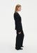 Side profile of a woman wearing a black long-sleeve top and matching wide-leg trousers, highlighting the clean lines and tailored fit of the outfit against a plain studio background.