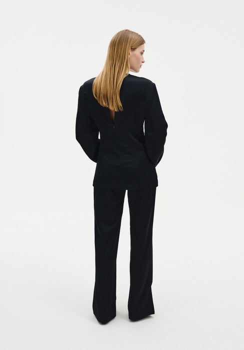Back view of a woman wearing a black long-sleeve top and matching tailored trousers, highlighting the clean silhouette and structured fit against a plain studio background.