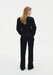 Back view of a woman wearing a black long-sleeve top and matching tailored trousers, highlighting the clean silhouette and structured fit against a plain studio background.