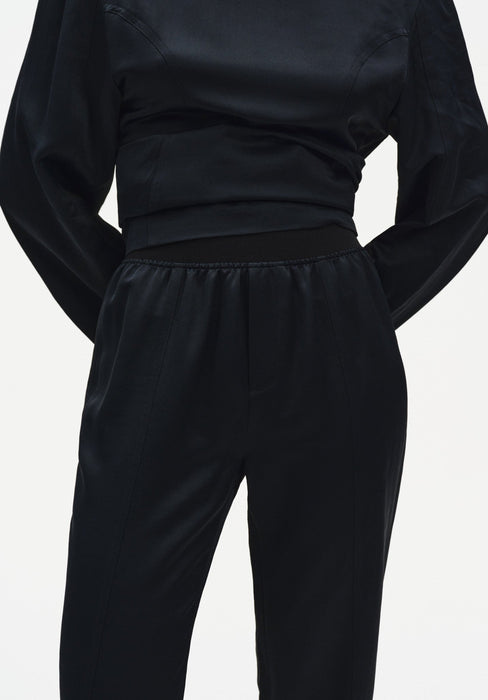 Close-up of a black outfit showing the back waist and upper trousers, featuring a smooth, slightly lustrous fabric, a wide fitted waistband, and softly gathered detailing at the top of the pants.