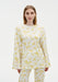 A woman stands facing forward against a plain white background, wearing a cream-colored long-sleeve top with a yellow floral pattern and matching trousers. The top has a relaxed fit with slightly flared sleeves, and she maintains a neutral expression.