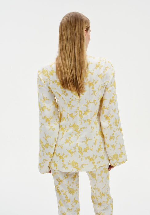 Rear view of a woman standing against a plain white background, wearing a cream-colored long-sleeve top with a yellow floral pattern and matching trousers. Her straight light brown hair falls down her back.