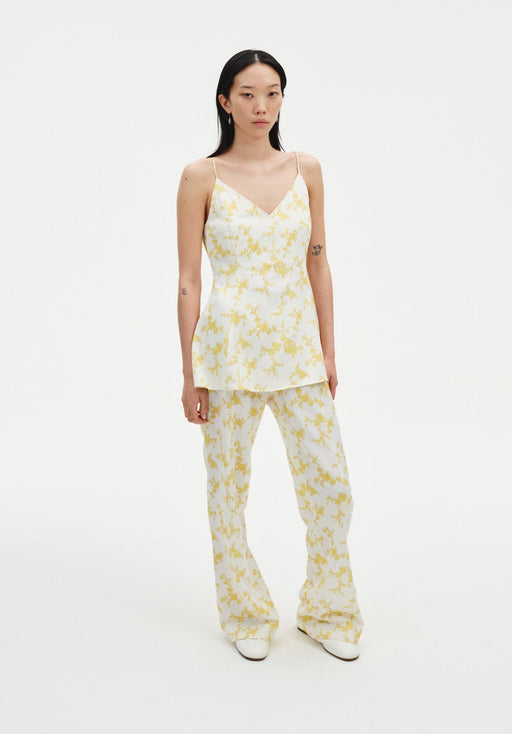 A woman with long dark hair stands against a plain background wearing a coordinated cream outfit with a yellow floral print, featuring a sleeveless V-neck top and matching wide-leg pants, paired with white shoes.