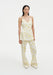 A woman with long dark hair stands against a plain background wearing a coordinated cream outfit with a yellow floral print, featuring a sleeveless V-neck top and matching wide-leg pants, paired with white shoes.