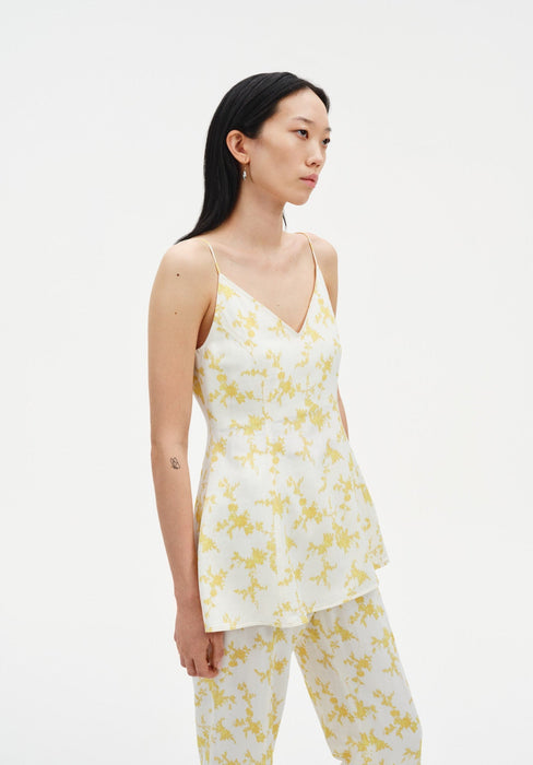 A woman with long dark hair is shown in a side-facing pose against a plain background, wearing a cream V-neck camisole with thin straps and a yellow floral print, paired with matching pants.