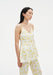 A woman with long dark hair is shown in a side-facing pose against a plain background, wearing a cream V-neck camisole with thin straps and a yellow floral print, paired with matching pants.