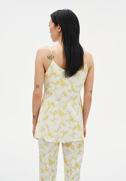 A woman with long dark hair is shown from the back against a plain background, wearing a cream camisole with thin straps and a yellow floral print, paired with matching pants.