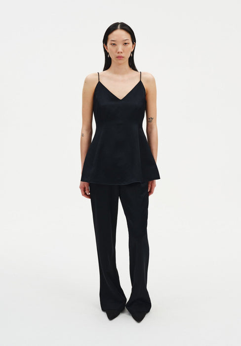 A woman stands facing forward against a plain white background, wearing a black sleeveless top with thin straps and a flared hem, paired with black trousers and pointed shoes. Her dark shoulder-length hair is worn down, and small tattoos are visible on her arms as she maintains a neutral expression.