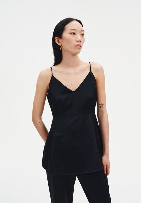 A woman stands in a three-quarter pose against a plain white background, wearing a black sleeveless top with thin straps and a softly flared fit, paired with black trousers. Her dark shoulder-length hair is worn down, and small tattoos are visible on her arms as she looks slightly to the side with a neutral expression.