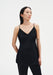 A woman stands in a three-quarter pose against a plain white background, wearing a black sleeveless top with thin straps and a softly flared fit, paired with black trousers. Her dark shoulder-length hair is worn down, and small tattoos are visible on her arms as she looks slightly to the side with a neutral expression.