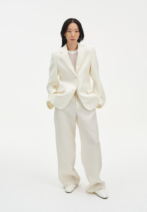 A woman stands facing forward against a plain white background, wearing a cream-colored tailored blazer with a single-button closure and matching wide-leg trousers, paired with light-colored shoes. Her dark shoulder-length hair is worn down, and she maintains a neutral expression with her hands in her pockets.