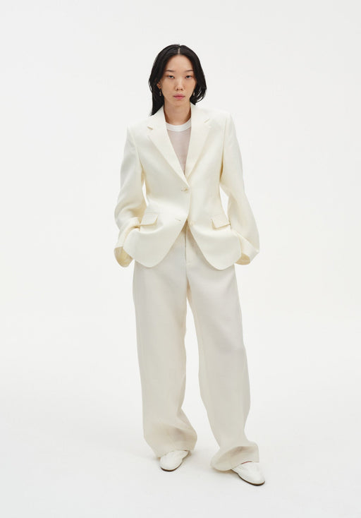 A woman stands facing forward against a plain white background, wearing a cream-colored tailored blazer with a single-button closure and matching wide-leg trousers, paired with light-colored shoes. Her dark shoulder-length hair is worn down, and she maintains a neutral expression with her hands in her pockets.