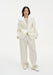 A woman stands facing forward against a plain white background, wearing a cream-colored tailored blazer with a single-button closure and matching wide-leg trousers, paired with light-colored shoes. Her dark shoulder-length hair is worn down, and she maintains a neutral expression with her hands in her pockets.
