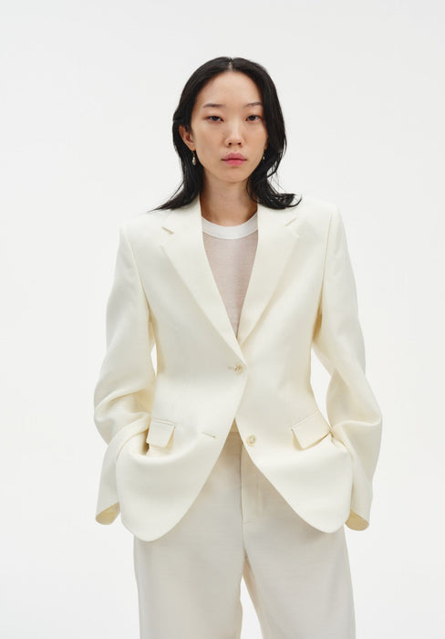 A woman faces forward against a plain white background, wearing a cream-colored tailored blazer with a single-button closure over a light top, paired with matching trousers. Her dark, shoulder-length hair is worn down, and she has a neutral expression with her hands in her pockets.