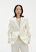 A woman faces forward against a plain white background, wearing a cream-colored tailored blazer with a single-button closure over a light top, paired with matching trousers. Her dark, shoulder-length hair is worn down, and she has a neutral expression with her hands in her pockets.
