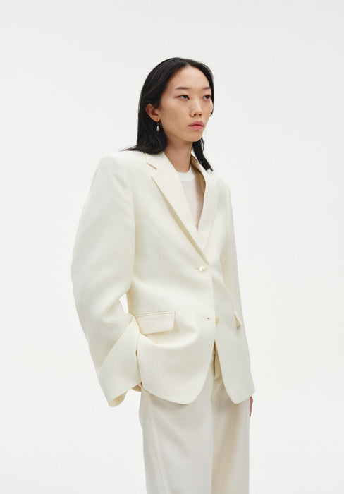 A woman stands in a three-quarter pose against a plain white background, wearing a cream-colored tailored blazer over a light top with matching trousers. Her dark shoulder-length hair is worn down, and she looks slightly to the side with a neutral expression, one hand resting in her pocket.