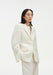 A woman stands in a three-quarter pose against a plain white background, wearing a cream-colored tailored blazer over a light top with matching trousers. Her dark shoulder-length hair is worn down, and she looks slightly to the side with a neutral expression, one hand resting in her pocket.