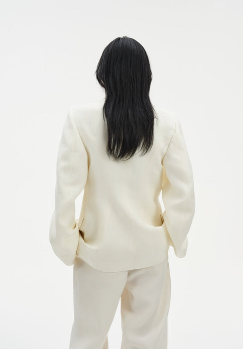 Rear view of a woman standing against a plain white background, wearing a cream-colored tailored blazer with structured shoulders and matching trousers. Her straight dark hair falls to mid-back, and her hands rest casually in her pockets.
