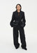 A woman stands facing forward against a plain white background, wearing a black tailored blazer over a sheer dark top with matching wide-leg trousers and black shoes. Her dark shoulder-length hair is worn down, and she maintains a neutral expression with her hands in her pockets.