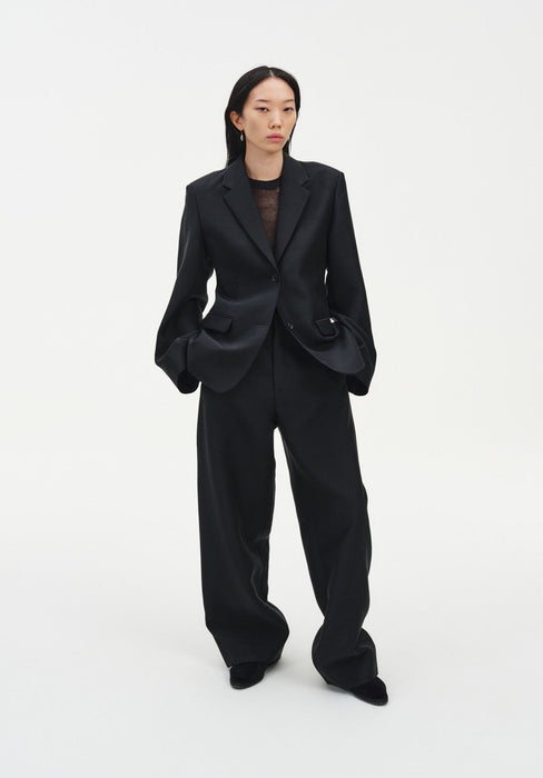 A woman stands facing forward against a plain white background, wearing a black tailored blazer over a sheer dark top with matching wide-leg trousers and black shoes. Her dark shoulder-length hair is worn down, and she has a neutral expression with her hands in her pockets.
