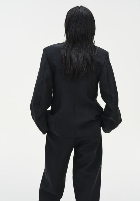 Rear view of a woman standing against a plain white background, wearing a black tailored blazer with structured seams and matching trousers. Her dark shoulder-length hair falls down her back, and her hands are placed in her pockets.