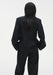 Rear view of a woman standing against a plain white background, wearing a black tailored blazer with structured seams and matching trousers. Her dark shoulder-length hair falls down her back, and her hands are placed in her pockets.