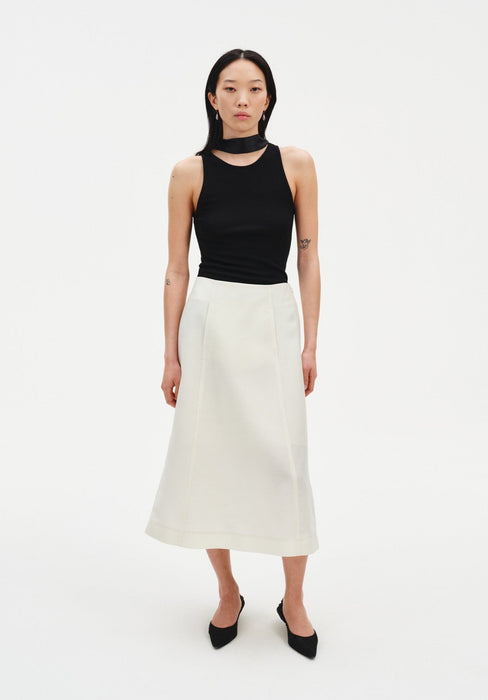 A woman stands facing forward against a plain white background, wearing a black sleeveless top with a high neckline and choker-style detail, paired with a cream-colored midi skirt and black pointed heels. Her dark shoulder-length hair is worn down, and she maintains a neutral expression with arms relaxed at her sides.