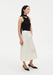 A woman stands in a three-quarter side pose against a plain white background, wearing a black sleeveless top with a high neckline and choker-style detail, paired with a cream-colored midi skirt and black pointed heels. Her dark shoulder-length hair is worn down, and she looks slightly to the side with a neutral expression.