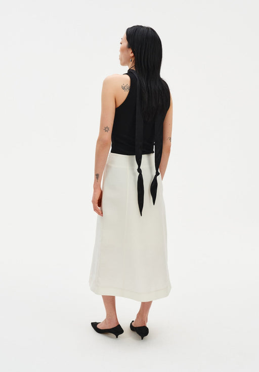 Rear three-quarter view of a woman against a plain white background, wearing a black sleeveless top with a long tie detail draping down the back, paired with a cream-colored midi skirt and black pointed heels. Her dark shoulder-length hair is worn down, and small tattoos are visible on her arms and upper back.